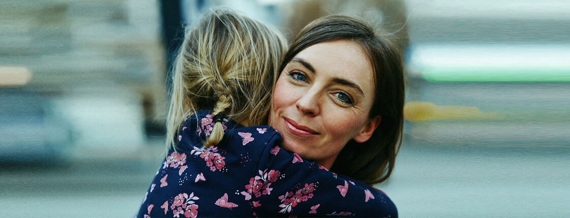 A woman in a floral blouse hugs a child and smiles warmly, while the background remains blurred