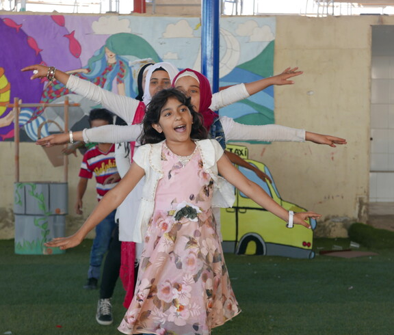 Girls with outstretched arms dance on a green floor in front of a colourful wall in the Bekaa Valley, Lebanon.