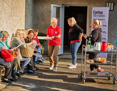 Several women are sitting on a bench discussing documents while a Caritas volunteer hands them paperwork. In the background is a table with drinks and food, as well as a Caritas banner that reads “Caritas: We Help.”
