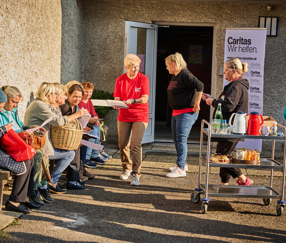 Several women are sitting on a bench discussing documents while a Caritas volunteer hands them paperwork. In the background is a table with drinks and food, as well as a Caritas banner that reads “Caritas: We Help.”
