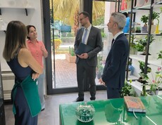 Four adults stand in a flower shop and are engaged in conversation; shelves with plants and decorations are visible in the room.
