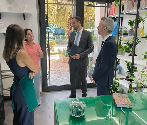 Four adults stand in a flower shop and are engaged in conversation; shelves with plants and decorations are visible in the room.