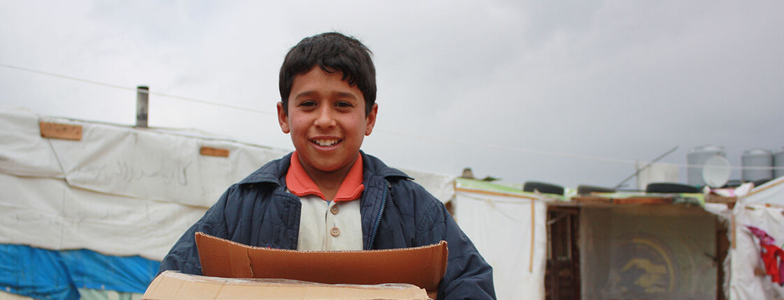 A refugee child is in a tent camp in Lebanon. He is holding a large box in his hands. 