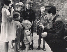 A man kneels in front of a group of children in the postwar period and talks to them—a scene in front of a wall, circa the 1940s