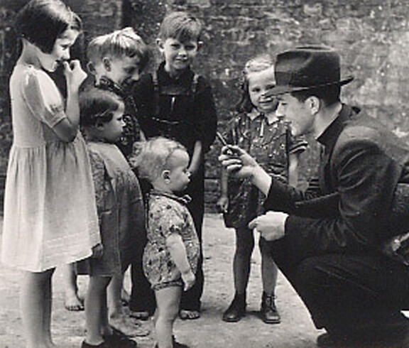 A man kneels in front of a group of children in the postwar period and talks to them—a scene in front of a wall, circa the 1940s