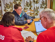 A woman is showing information on her smartphone while sitting at a table with others. The group is discussing aid for flood victims in Böheimkirchen. A laptop and a passport are visible on the table, suggesting that this is a formal consultation.