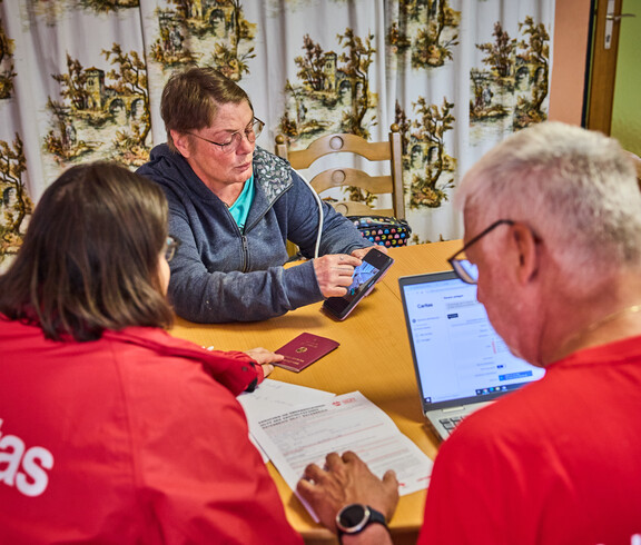 A woman is showing information on her smartphone while sitting at a table with others. The group is discussing aid for flood victims in Böheimkirchen. A laptop and a passport are visible on the table, suggesting that this is a formal consultation.
