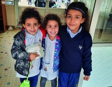 Three girls in school uniforms stand smiling in front of the St. Vincent School building in Broumana, Lebanon.