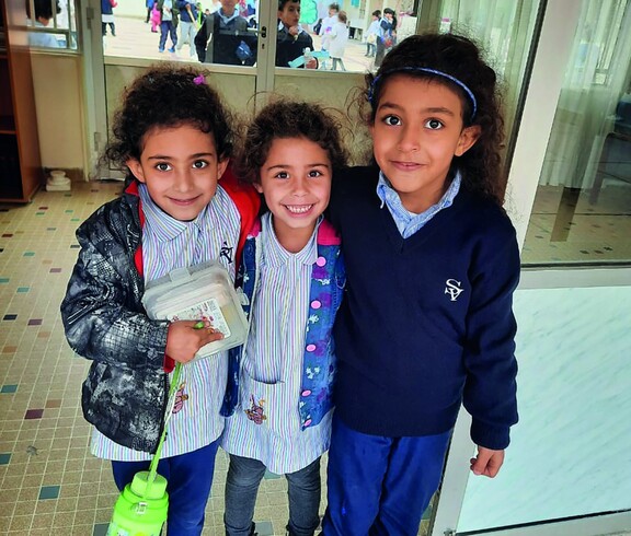 Three girls in school uniforms stand smiling in front of the St. Vincent School building in Broumana, Lebanon.