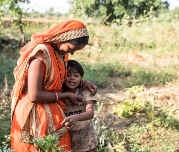 A small-scale farmer in traditional clothing stands with her child in a field, holding them lovingly.
