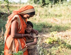 A small-scale farmer in traditional clothing stands with her child in a field, holding them lovingly.