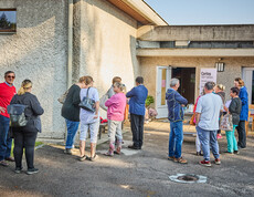 People are gathering in front of a building in Böheimkirchen to receive emergency flood relief from Caritas. A sign reading “Caritas: We Help” is visible as the people stand in a loose group and talk to one another.