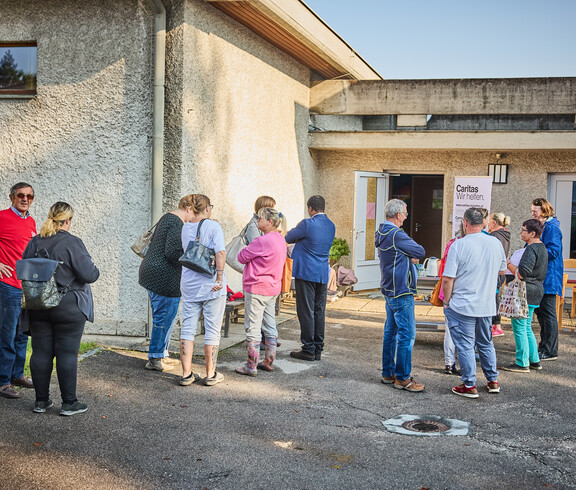 People are gathering in front of a building in Böheimkirchen to receive emergency flood relief from Caritas. A sign reading “Caritas: We Help” is visible as the people stand in a loose group and talk to one another.