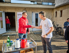 Two men are shaking hands in front of a building where Caritas is organizing emergency aid. One is wearing a red Caritas jacket, while the other is wearing a white T-shirt. Other people and a Caritas banner are visible in the background.
