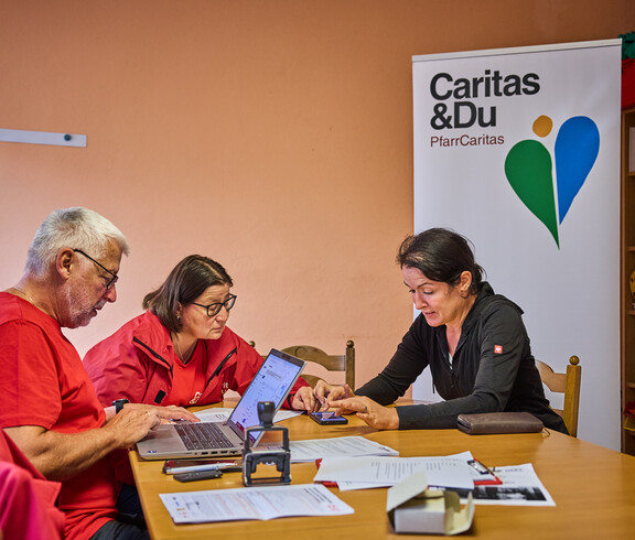 Three people are sitting at a table discussing emergency relief measures for flood victims in Böheimkirchen. Two of them are wearing red Caritas jackets, while a woman is pointing to a document. A Caritas banner is visible in the background.