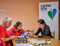Three people are sitting at a table discussing emergency relief measures for flood victims in Böheimkirchen. Two of them are wearing red Caritas jackets, while a woman is pointing to a document. A Caritas banner is visible in the background.