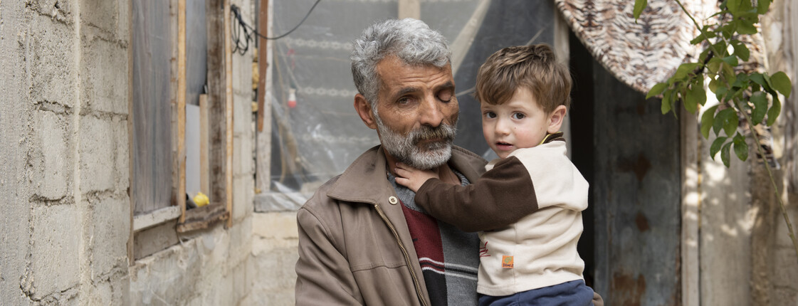 Elderly man holding a small child in front of a damaged house in Syria.