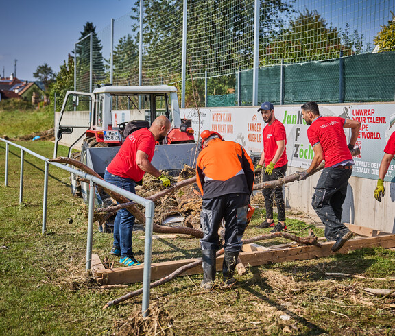 Volunteers are clearing tree trunks and debris from a sports field in Hofstetten-Grünau. They are wearing red Caritas T-shirts, and in the background, a tractor stands by, ready to haul away the debris. The volunteers are working together to repair the flood damage.