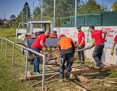 Volunteers are clearing tree trunks and debris from a sports field in Hofstetten-Grünau. They are wearing red Caritas T-shirts, and in the background, a tractor stands by, ready to haul away the debris. The volunteers are working together to repair the flood damage.