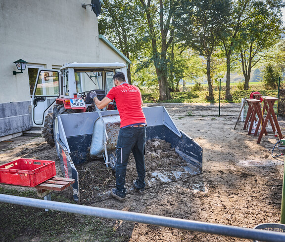 A volunteer is shoveling mud into a tractor bucket in front of a building in Hofstetten-Grünau. The man is wearing a red T-shirt that reads “We Help.” In the background, you can see trees and tools being used for the cleanup efforts.