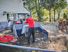 A volunteer is shoveling mud into a tractor bucket in front of a building in Hofstetten-Grünau. The man is wearing a red T-shirt that reads “We Help.” In the background, you can see trees and tools being used for the cleanup efforts.