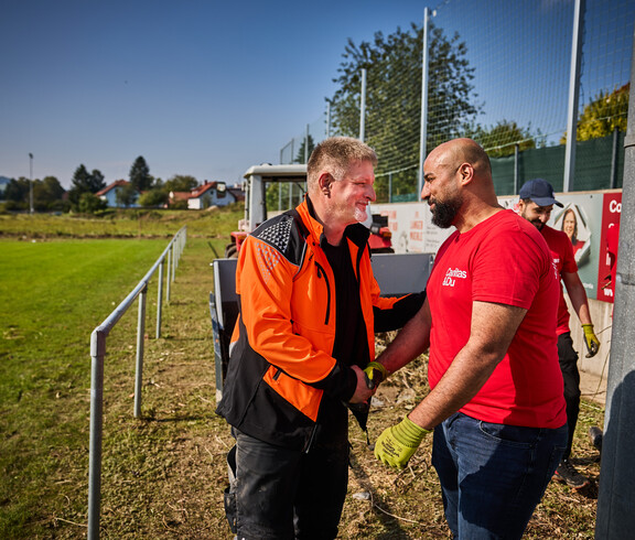 A man in an orange work jacket smiles as he shakes hands with a volunteer wearing a red Caritas T-shirt. In the background, a tractor and a sports field are visible, where other volunteers are helping with the cleanup efforts.