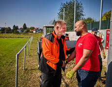 A man in an orange work jacket smiles as he shakes hands with a volunteer wearing a red Caritas T-shirt. In the background, a tractor and a sports field are visible, where other volunteers are helping with the cleanup efforts.