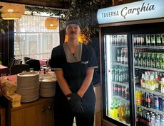 A young man in work attire stands in a restaurant next to a refrigerator with drinks and stacked dishes.