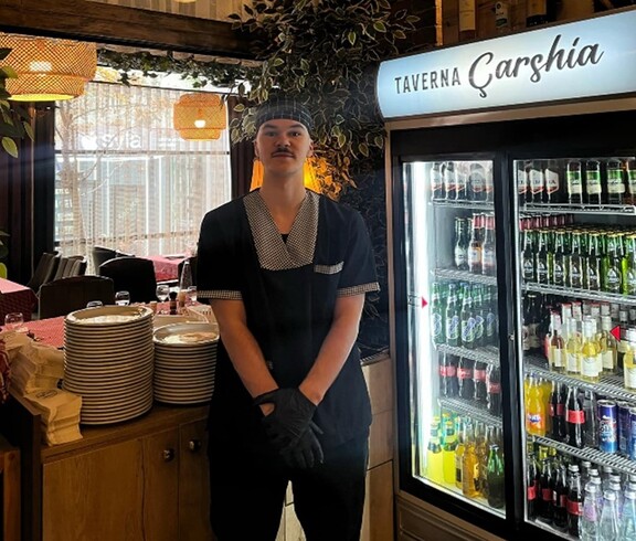 A young man in work attire stands in a restaurant next to a refrigerator with drinks and stacked dishes.