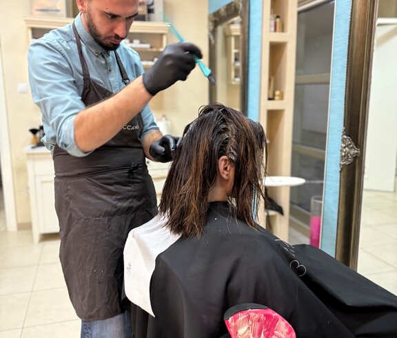 A person is applying hair color to a seated individual. In the foreground, a color bowl and work tools are visible.