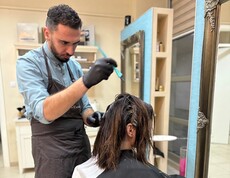 A person is applying hair color to a seated individual. In the foreground, a color bowl and work tools are visible.