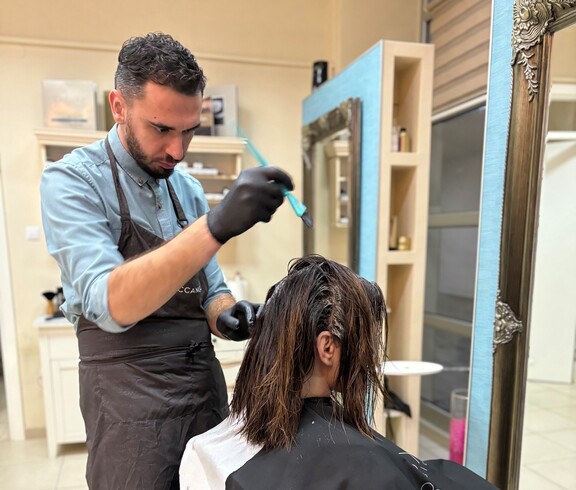 A person is applying hair color to a seated individual. In the foreground, a color bowl and work tools are visible.