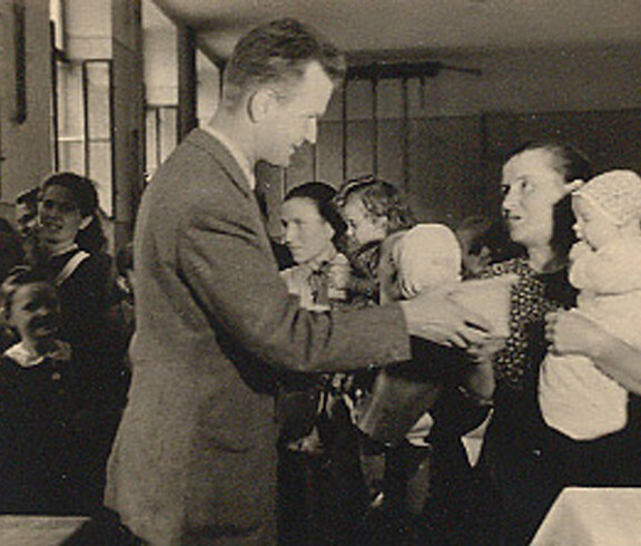 A man hands a package of sugar to a mother with a child—a scene from a Caritas food distribution in 1946