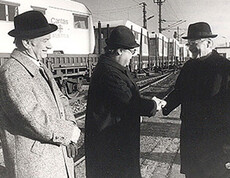Three men are standing in front of a Caritas relief train and shaking hands—in the background, relief cars bearing the words “Caritas Austria,” Southern Italy, 1980