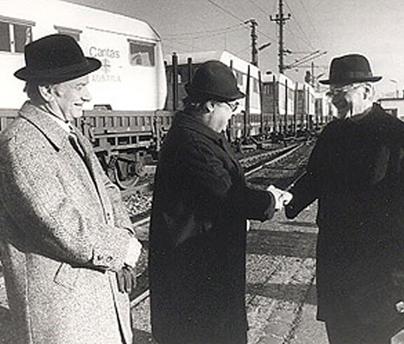 Three men are standing in front of a Caritas relief train and shaking hands—in the background, relief cars bearing the words “Caritas Austria,” Southern Italy, 1980