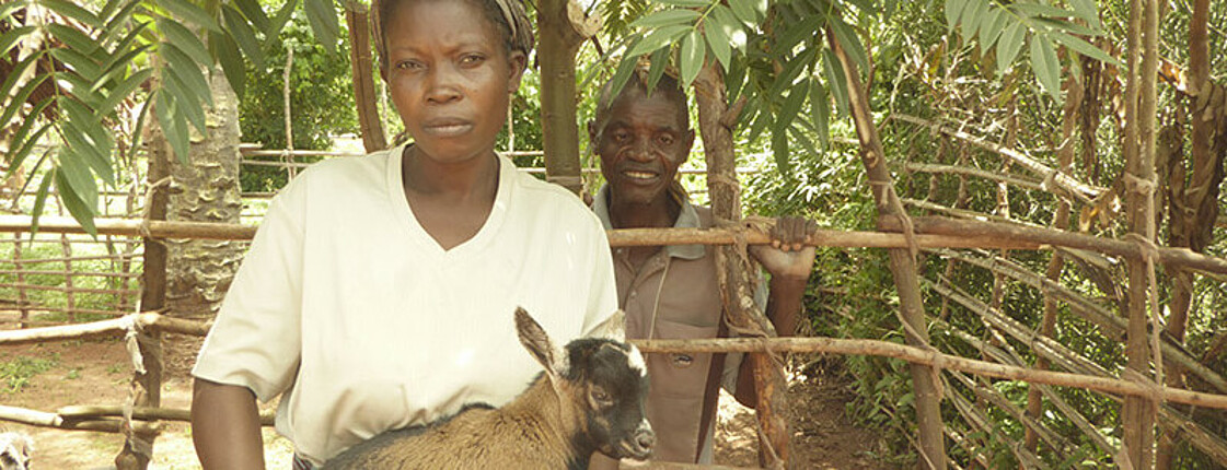 Woman holding a goat, in the background a man smiling at the camera